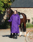 EQUIDRY women's long waterproof horse riding coat in deep purple/grey modelled by rider stood with horse 