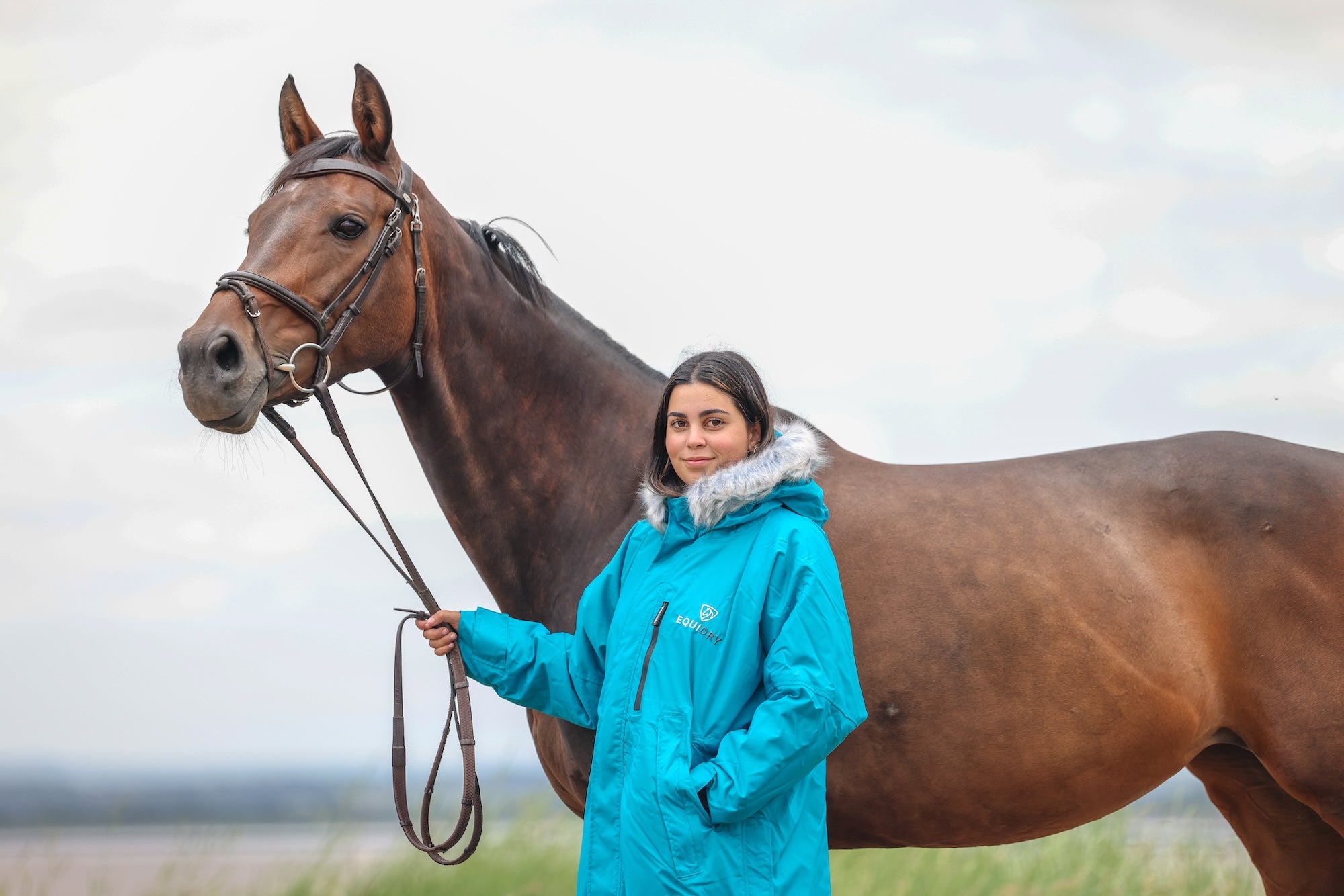 EQUIDRY Evolution Lux Women's Warm Waterproof Winter Horse Riding Coat Thick Fleece Lining with Faux Fur Trim Hood in turquoise rider stood with horse