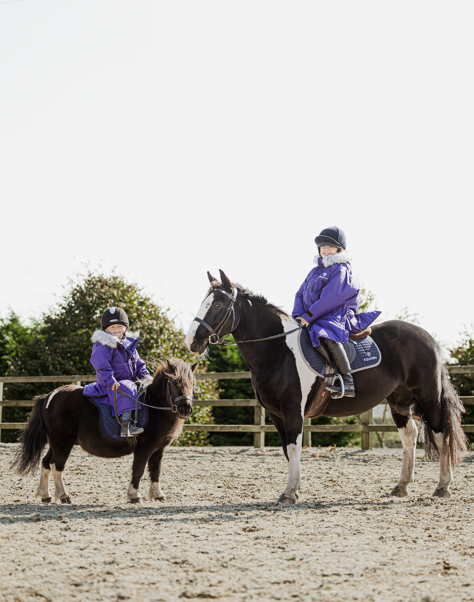 EQUIDRY Evolution Lux children's Warm Waterproof Winter Horse Riding Coat Thick Fleece Lining with Faux Fur Trim Hood being shown in purple by two kids riding their horses 