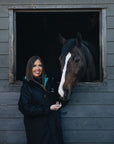 EQUIDRY women's long waterproof horse riding coat in black/turquoise modelled by rider on yard with horse 