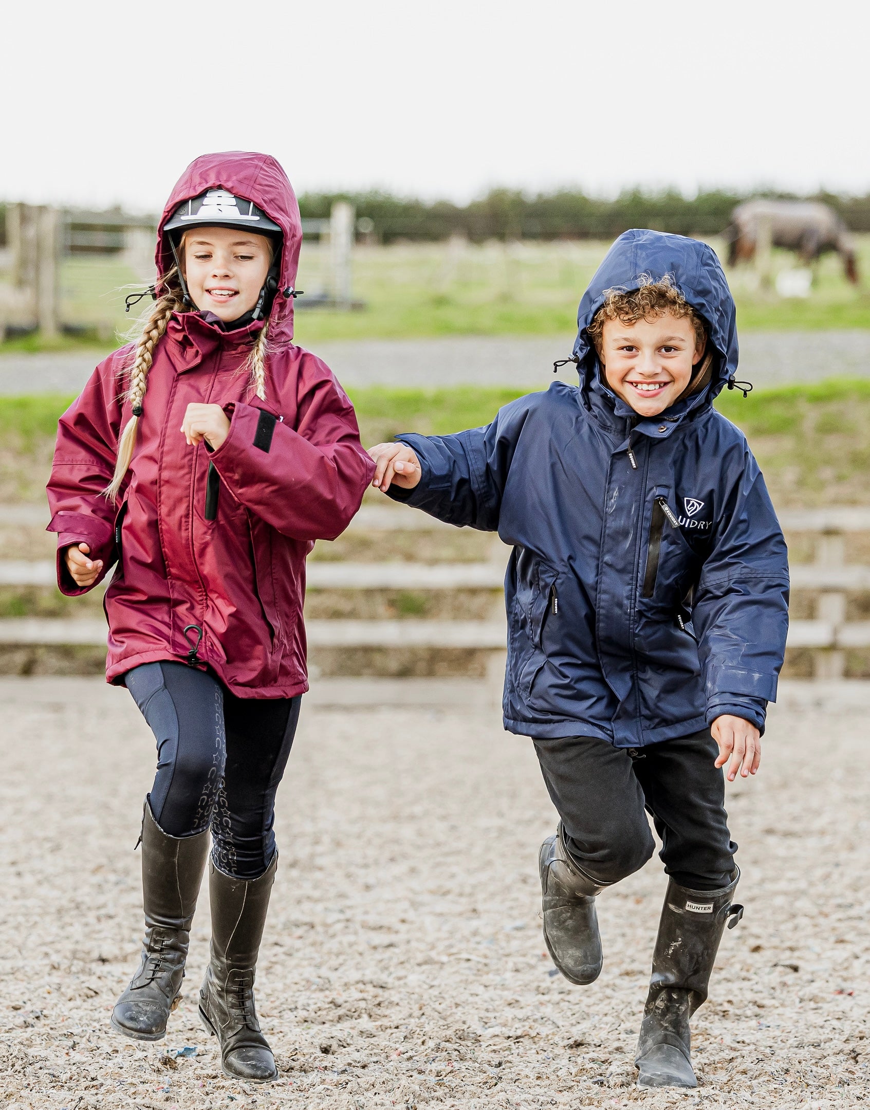 Shortie Child's Oversized Short Waterproof blouson style Riding Jacket Thin Fleece Lining Stowaway Hood in plum and navy worn by kids running in arena 