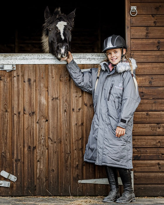 EQUIDRY Evolution Lux children's Warm Waterproof Winter Horse Riding Coat Thick Fleece Lining with Faux Fur Trim Hood being shown in charcoal/pale pink by girl with horse on the yard