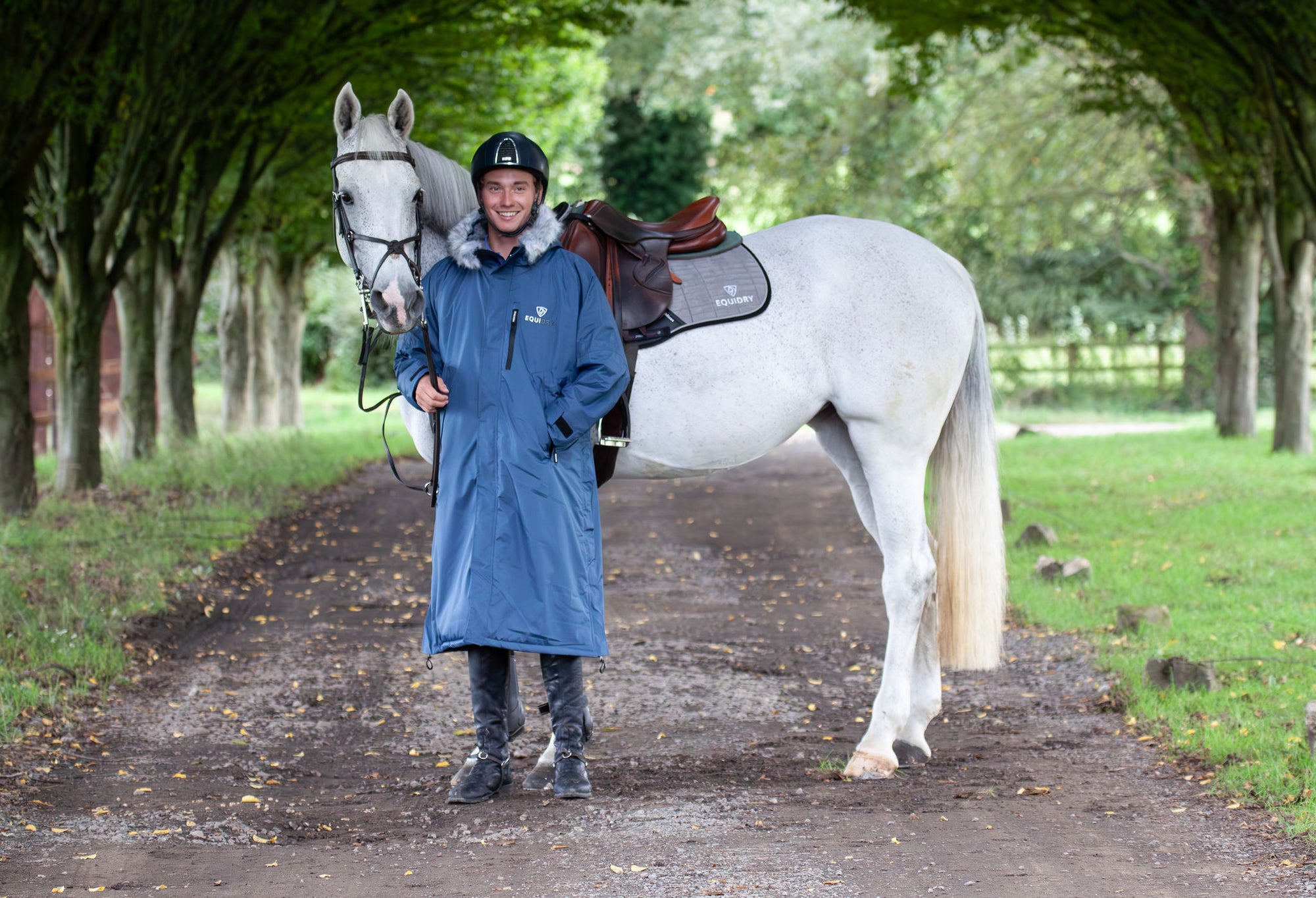 Evolution Lux Men&#39;s Warm Waterproof Horse Riding Coat in steel blue worn by male rider stood with horse 