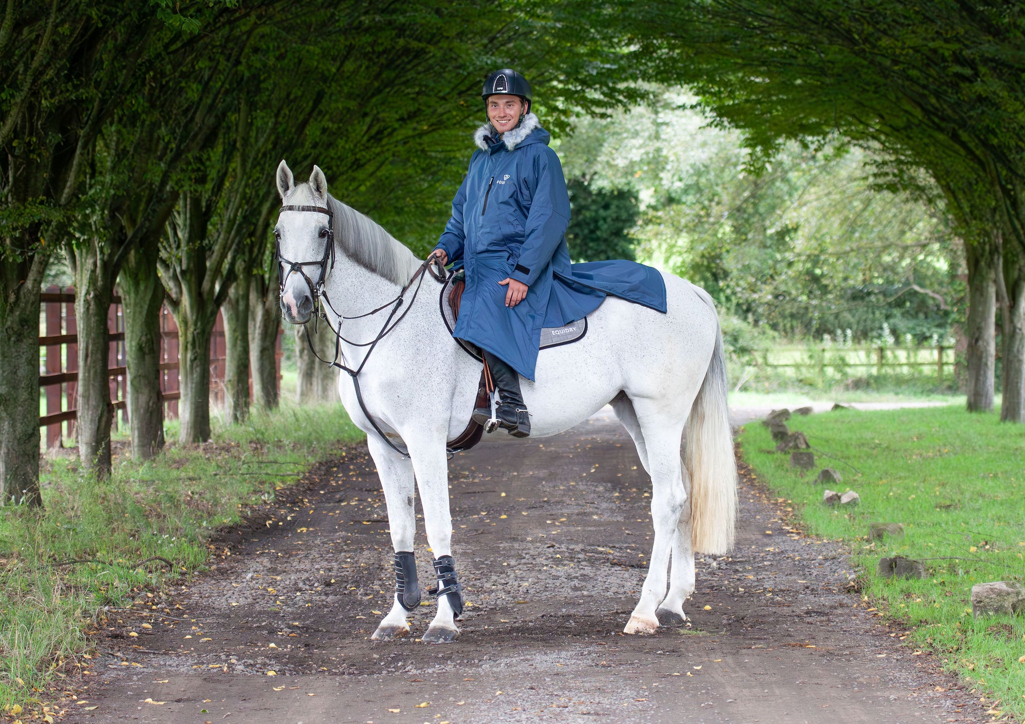 Evolution Lux Men&#39;s Warm Waterproof Horse Riding Coat in steel blue modelled by male rider on horseback showing storm skirts and side zips