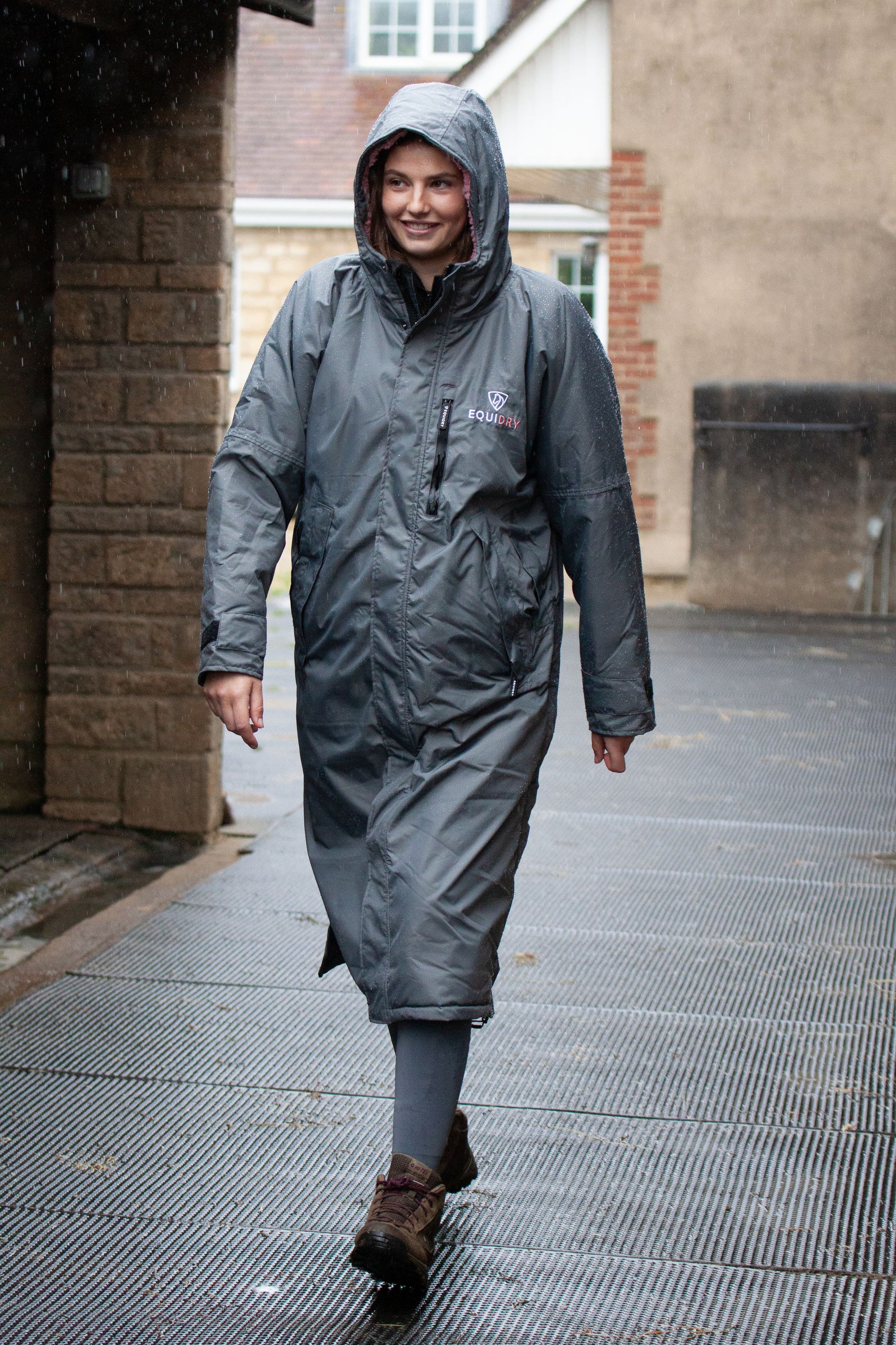 EQUIDRY women's long waterproof horse riding coat in charcoal/pale pink showing groom on yard sheltering from the rain 