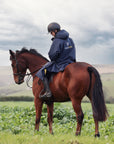 EQUIDRY women's long waterproof horse riding coat in navy/yellow modelled by rider hacking on horse through fields 