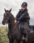 EQUIDRY women's long waterproof horse riding coat in black/olive modelled by rider out hacking, showing storm skirts and side zips